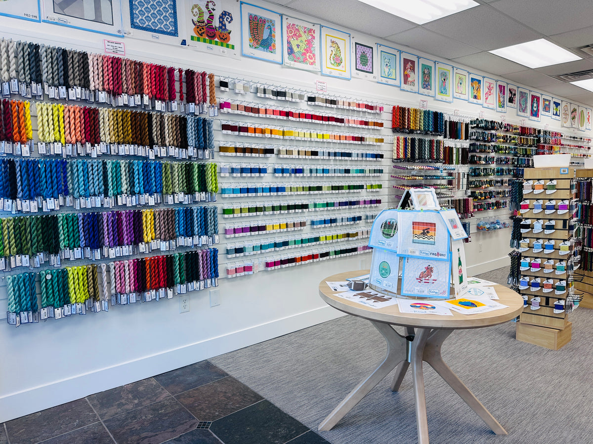 A large display of colorful embroidery floss or thread spools mounted on wall racks inside a craft supply store. A small round table in the foreground holds a light blue quilted display bag with embroidered text and illustrations.