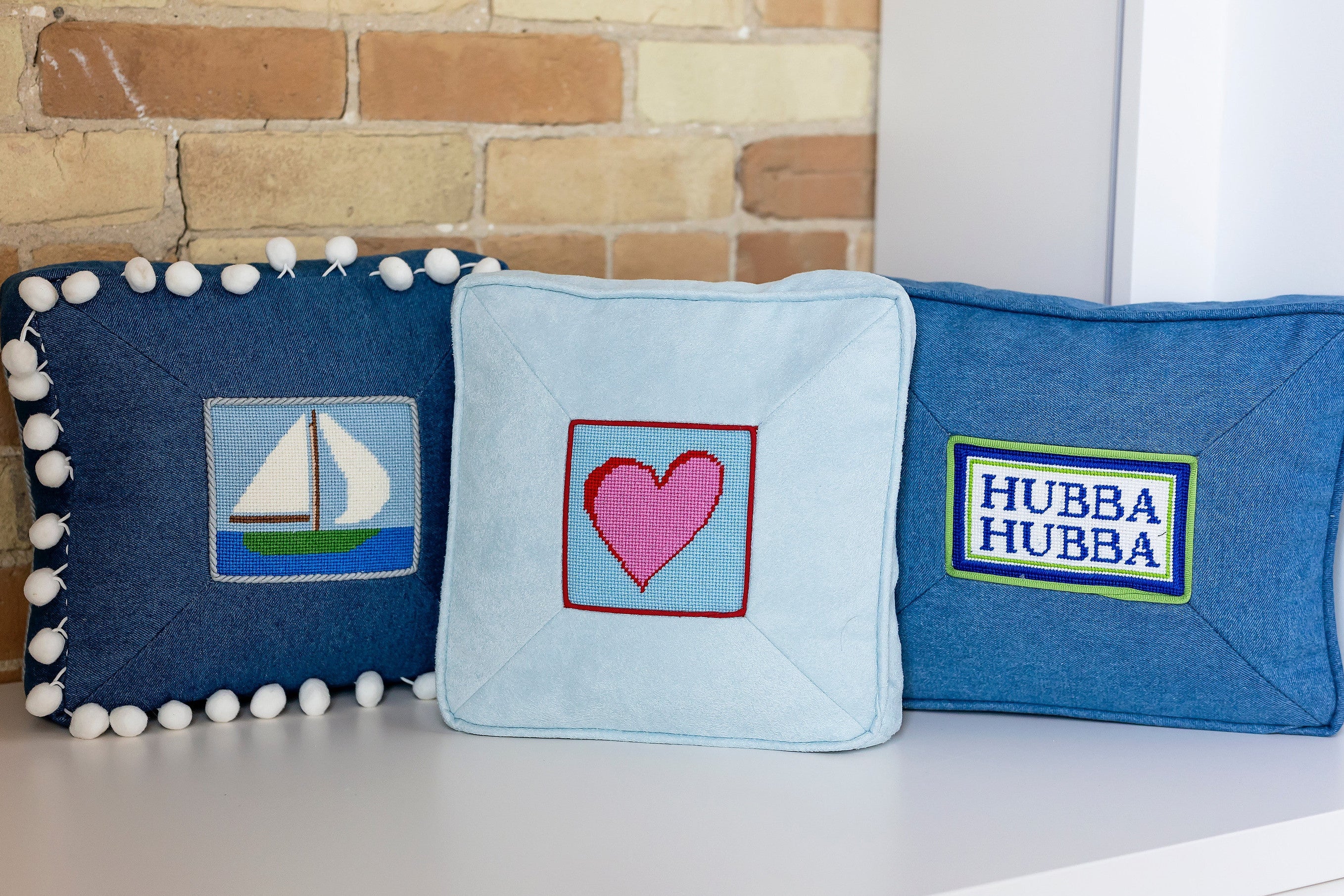 Three decorative pillows with needlepoint designs on a white surface against a brick wall.