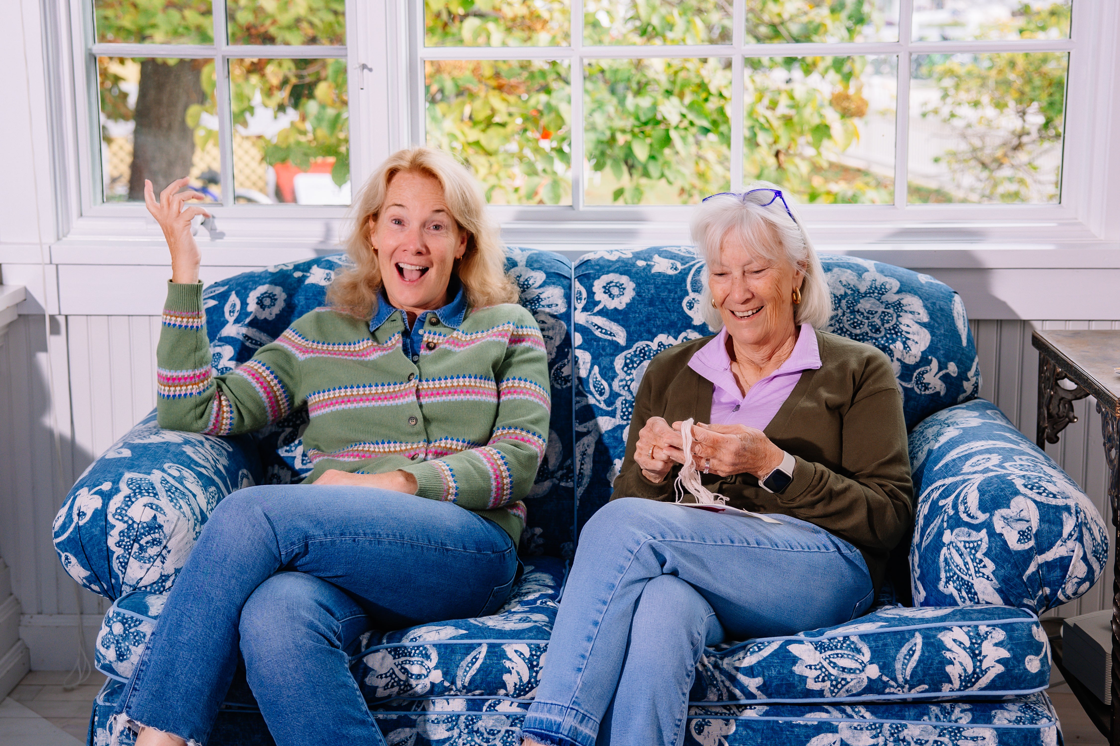 Two women sitting on a blue floral-patterned couch in a sunroom.
