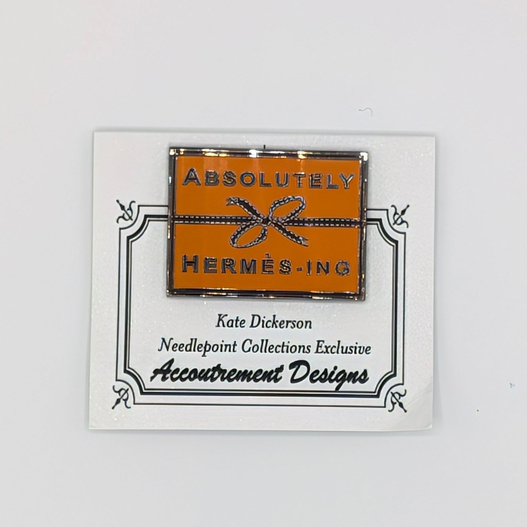 A small rectangular orange and black Hermès pin mounted on a white display card, photographed against a plain white background.