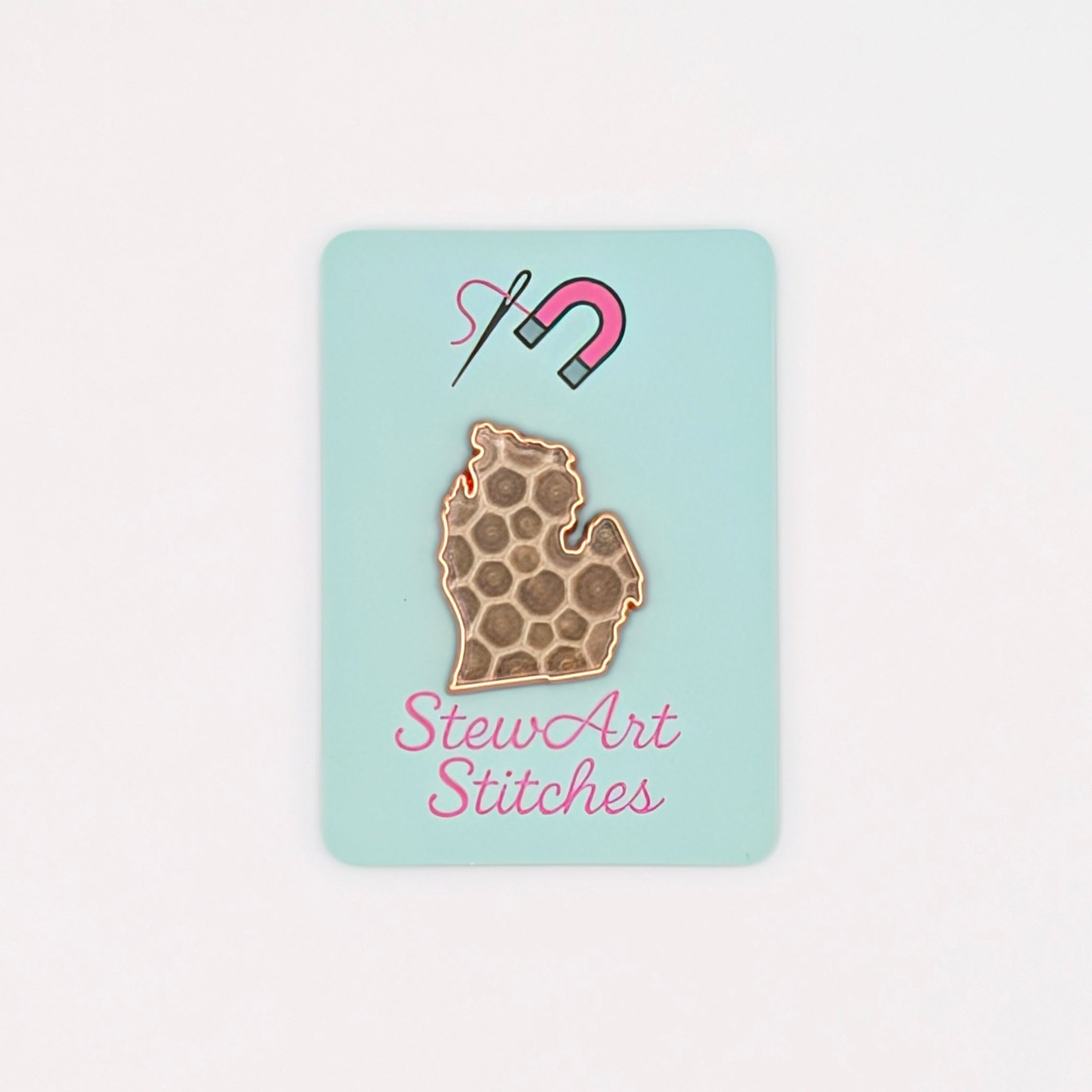 A small rectangular mint card featuring a brown patterned mitten with a pink and blue magnet and the words 'StewArt Stitches' in pink, centered on a plain white background.