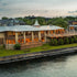 A large waterfront building with a stone and wood facade, warm glowing windows, and a metal roof, situated beside calm water with trees and other structures in the background at dusk.